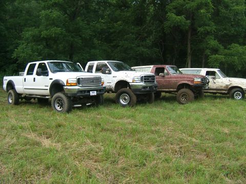 1993 Ford Bronco - A lineup of four lifted pickup trucks, including a maroon model and three white ones, are parked side-by-side in a grassy outdoor setting.
