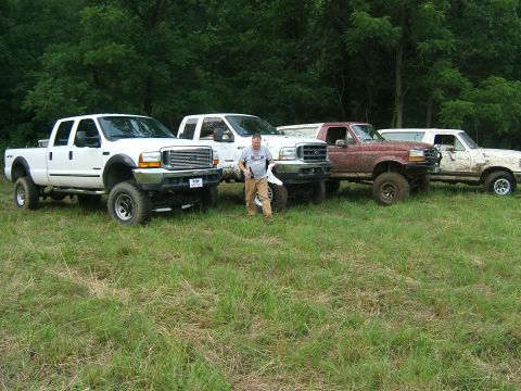 1993 Ford Bronco - Several lifted Ford trucks are parked in a grassy area with a wooded background.