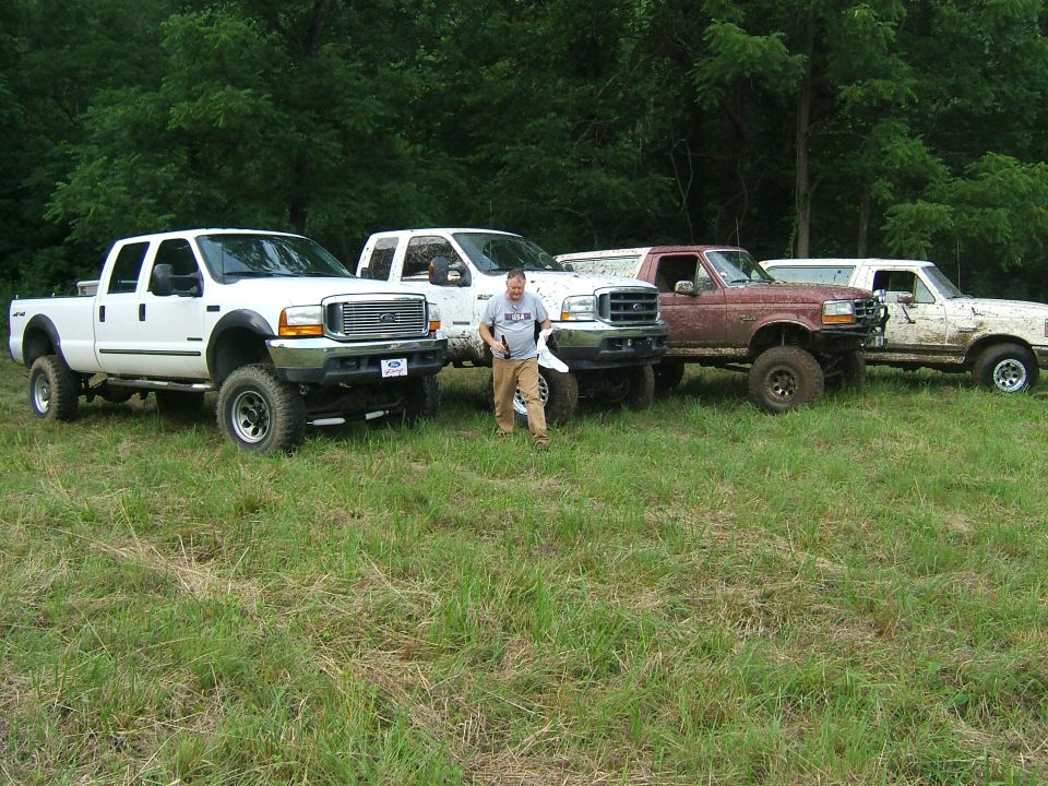 Four lifted Ford F-Series trucks parked in a grassy field with trees in the background. The trucks appear to be from the late 1990s
