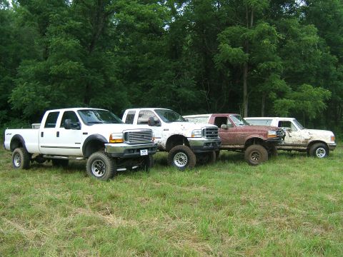 1993 Ford Bronco - A lineup of four lifted pickup trucks, some covered in mud, are parked in a grassy area with a wooded background.