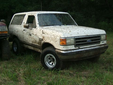 1993 Ford Bronco - Mud-covered white Ford Bronco with aftermarket wheels sits in a grassy area next to another vehicle.