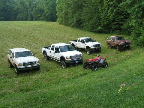 1993 Ford Bronco - A collection of four-wheel drive vehicles, including a 1993 Ford Bronco, are parked on a mowed grassy slope with trees in the background.