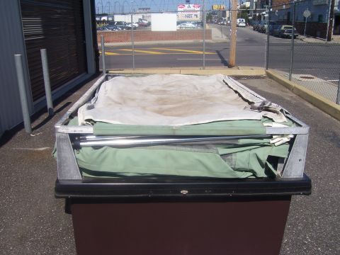 1991 Ford Bronco - A removed pale-green canvas soft top with its aluminum bow frame and chrome tube, folded and laid across the lid of a brown dumpster in an…