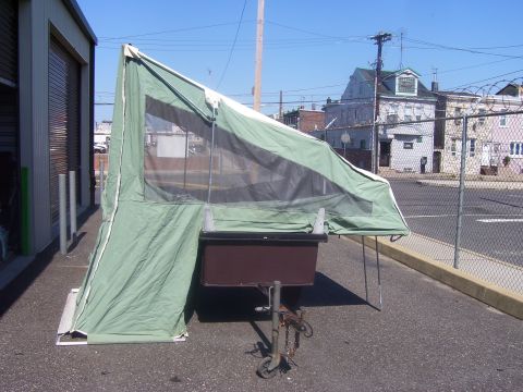 1991 Ford Bronco - A small tent camper trailer with a green and white canvas pop-up tent partially set up over a brown storage box body, parked on pavement be…