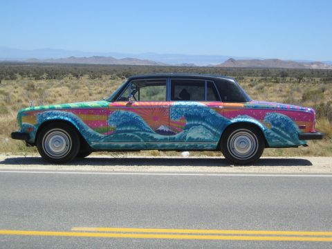 1968 Ford Mustang - A car with a vibrant mural depicting oceanic waves and a sunrise is parked beside a desert highway.