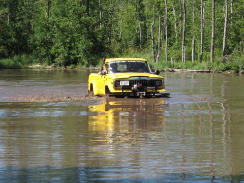 1977 Ford F-150 - This yellow Jeep pickup truck is navigating a shallow water crossing, kicking up mud as it goes.