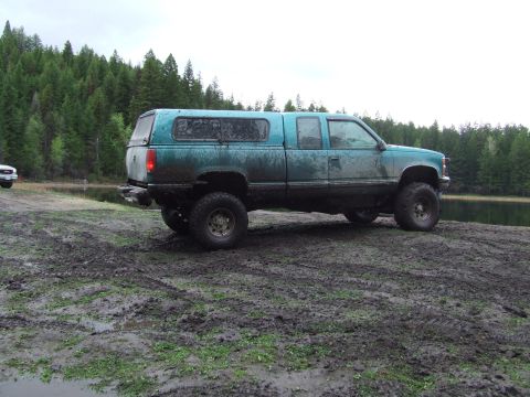 1990 Ford Bronco - A muddy teal pickup truck with a camper shell sits by a lake, its tires leaving tracks in the wet ground.