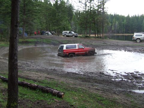 1990 Ford Bronco - Several vehicles are parked near a campsite by a lake, while one SUV navigates a muddy puddle.