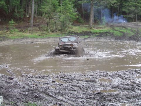 1990 Ford Bronco - A Jeep Wrangler navigates a muddy obstacle, kicking up water and mud.