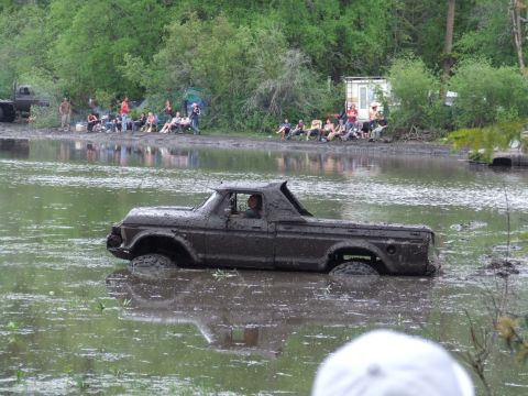 1990 Ford Bronco - A mud-covered pickup truck navigates through a muddy body of water with spectators watching from the bank.