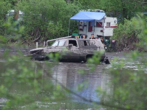1990 Ford Bronco - A heavily mud-covered SUV is partially submerged in water, with onlookers gathered on the bank behind it.
