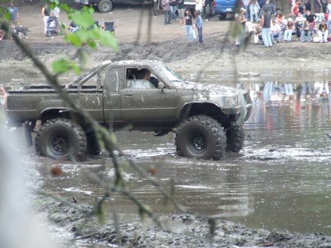 1990 Ford Bronco - A heavily muddied pickup truck navigates through a water obstacle, with spectators gathered on the bank.