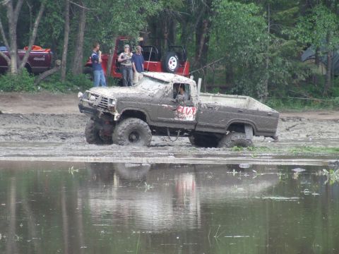 1990 Ford Bronco - Muddy pickup truck stuck in a mud pit with people on top. A red Jeep is visible behind.
