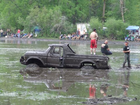 1990 Ford Bronco - A mud-covered pickup truck is stuck in a muddy, watery area while spectators watch from the bank.