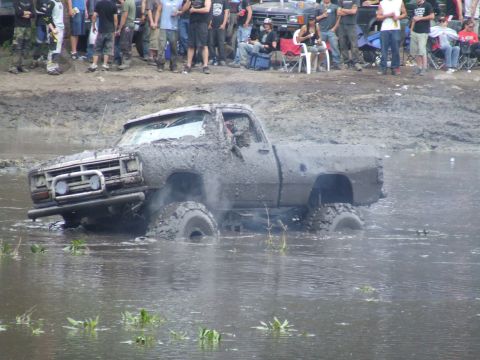 1990 Ford Bronco - A heavily mud-covered pickup truck is stuck in murky water with spectators in the background.