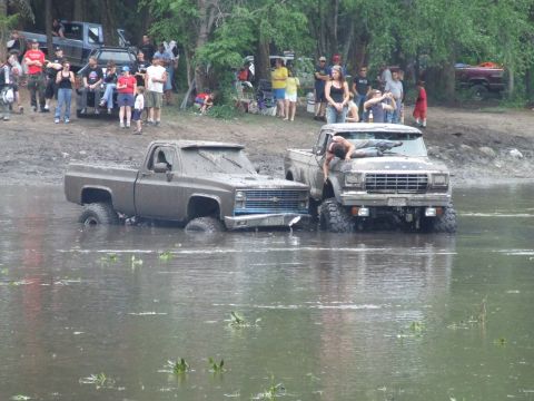 1990 Ford Bronco - Two heavily muddied pickup trucks are stuck in shallow water during an outdoor event, with onlookers gathered on the bank.