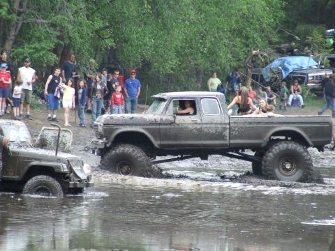 1990 Ford Bronco - Two off-road vehicles are stuck in the mud while spectators watch from the bank.