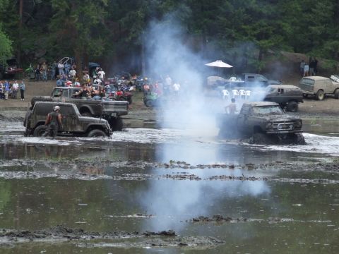 1990 Ford Bronco - Off-road vehicles churn through mud and create smoke during a weekend event, with onlookers gathered to watch the side.