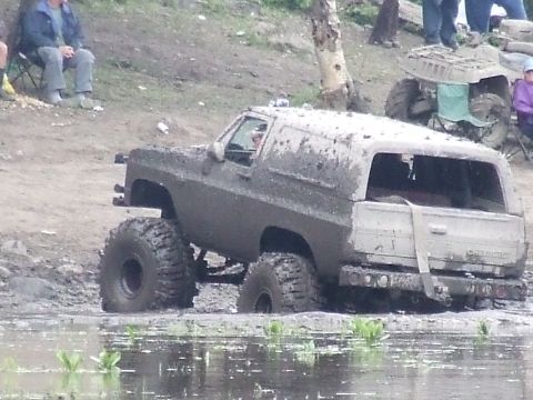 1990 Ford Bronco - This heavily modified SUV is deep in the mud and water, with spectators and another vehicle nearby.