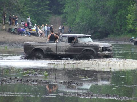 1990 Ford Bronco - A muddy Ford pickup truck navigates through a water obstacle during an outdoor event.
