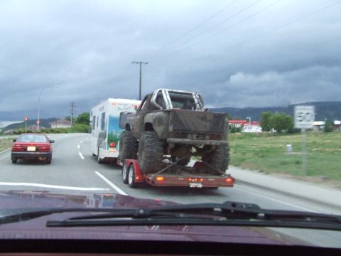 1990 Ford Bronco - A heavily modified, mud-covered truck is being transported on a trailer, followed by an RV on a cloudy day.