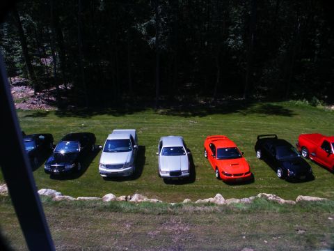 2001 Mercury Grand Marquis - Multiple vehicles are parked on a lawn in front of a wooded area.