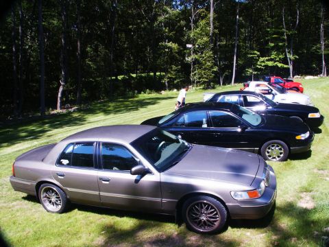 2001 Mercury Grand Marquis - A row of sedans and a truck are parked on a grassy lawn in front of a wooded area.