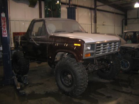 1982 Ford Bronco - A brown and tan two-tone 1980s Ford Bronco lifted on oversized mud-terrain tires with black steel wheels, parked in a garage next to a two-…