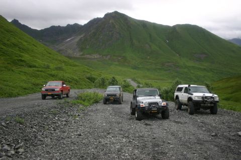 2003 Jeep TJ - Off-road vehicles travel on a gravel road through a scenic mountain pass under a cloudy sky.