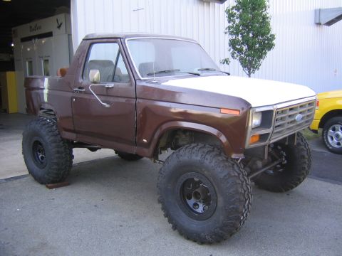 1982 Ford Bronco - A lifted Ford Bronco with oversized tires sits outside a garage, with a yellow vehicle partially visible to the right.