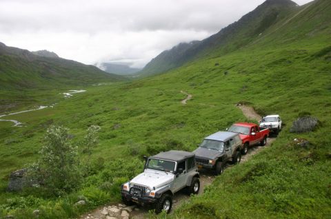 2003 Jeep TJ - A line of off-road vehicles navigates a winding path through a lush, green mountain landscape under a cloudy sky.