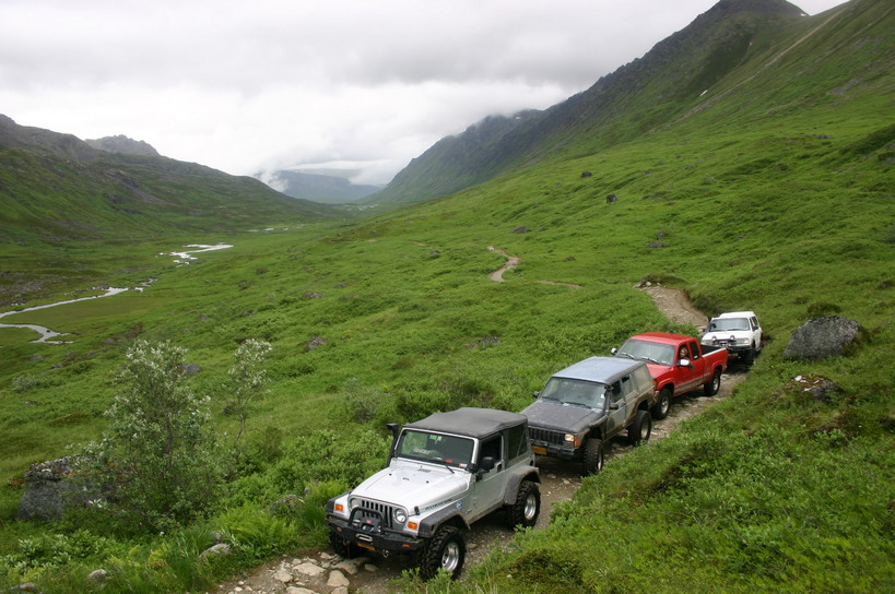 Four-door Jeep Wrangler, red pickup truck, gray SUV, and white SUV driving on a rocky trail through a green mountain valley