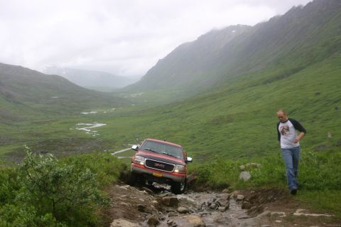2003 Jeep TJ - A red GMC pickup truck drives off-road through a rocky stream bed in a mountainous, green valley.