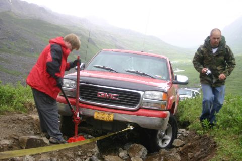 2003 Jeep TJ - Two people work to free a red GMC pickup truck stuck on a rough, rocky trail in a mountainous, overcast landscape.
