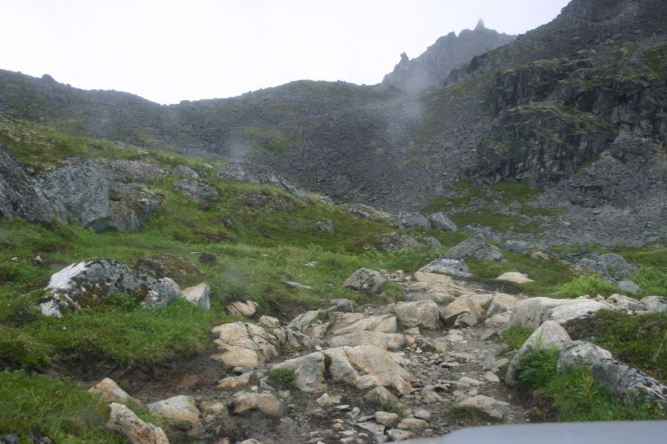 Rocky, grassy mountain slope with a rough, rocky path leading upwards. Fog or mist is visible in the background