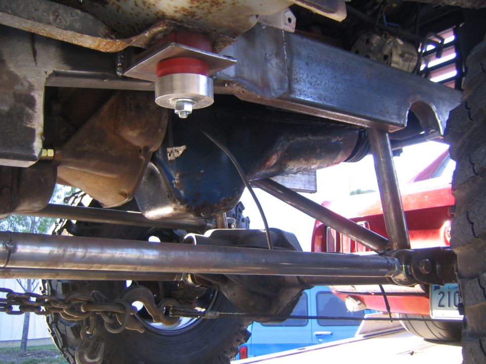 Underbody view of a vehicle with aftermarket suspension components, including a red and silver bushing mount. A chain and hook are visible near the tire