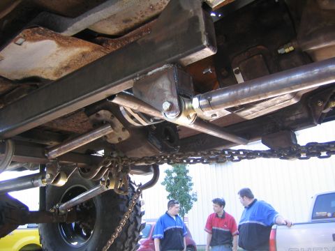 1982 Ford Bronco - The underside of a heavily modified vehicle shows custom suspension components and large chains, with three men observing.