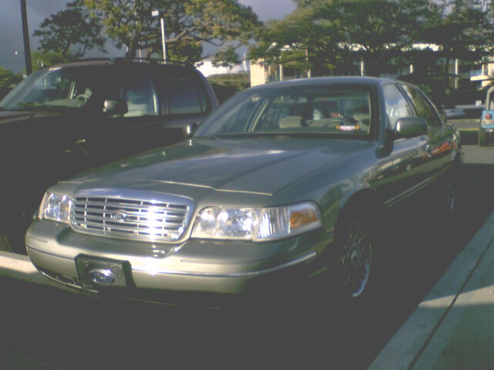 Sage green Ford Crown Victoria sedan, front three-quarter view, parked in a lot with trees behind