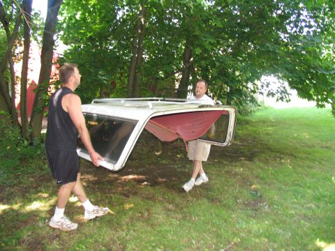 1986 Ford C-350 (Centurion Conversions) - Two men carry a detached car top outdoors near trees and grass.