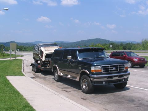 1986 Ford C-350 (Centurion Conversions) - A dark green Ford Bronco tows a trailer carrying a camper shell down a highway with a red SUV in the background.