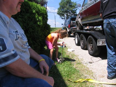 1986 Ford C-350 (Centurion Conversions) - A girl works on equipment near a trailer carrying a vehicle, with another truck visible in the background.