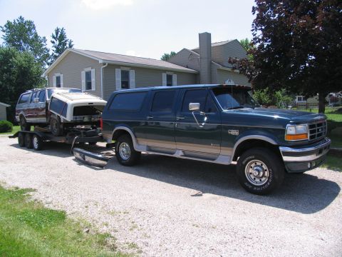 1986 Ford C-350 (Centurion Conversions) - A green Ford Bronco Centurion tows a trailer carrying a disassembled Ford Bronco on a gravel driveway.