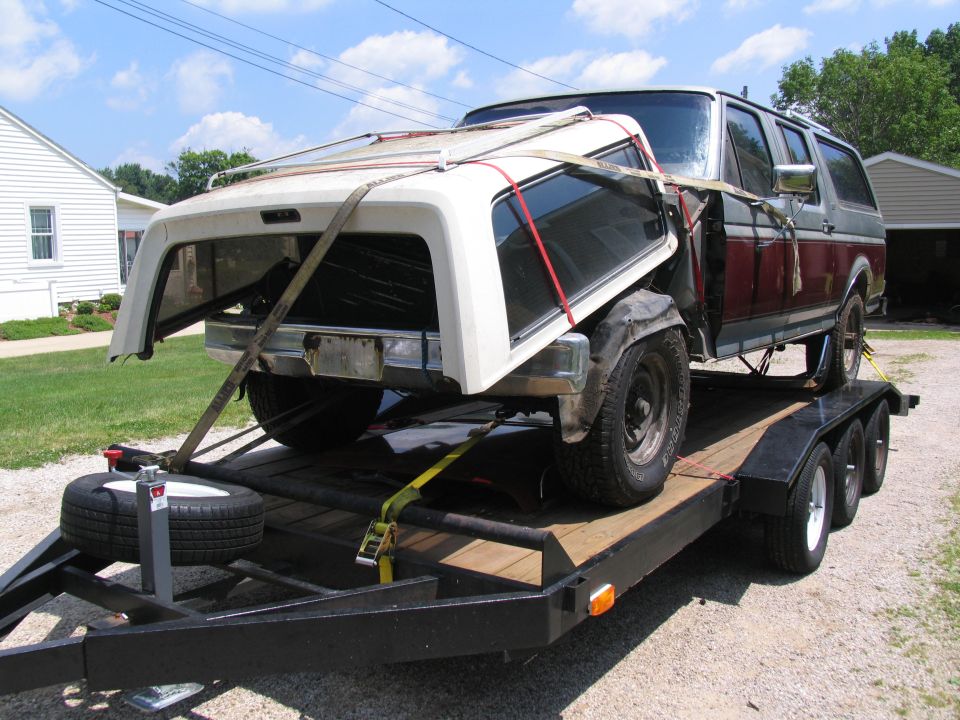 Maroon and silver Ford SUV body strapped to a black triple-axle trailer with white cab shell