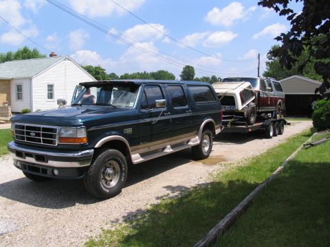 1986 Ford C-350 (Centurion Conversions) - A dark green Ford SUV tows a trailer carrying another SUV on a gravel driveway.