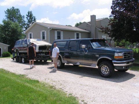 1986 Ford C-350 (Centurion Conversions) - A dark green Ford SUV conversion is parked on a gravel driveway, towing a trailer with a white camper shell.
