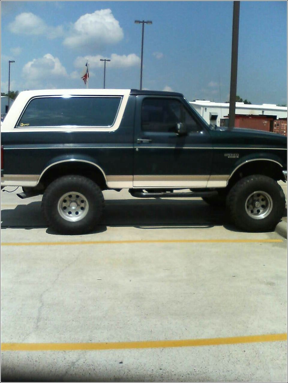 Dark green and tan 1992 Ford Bronco SUV, side view, lifted with large tires, white removable top
