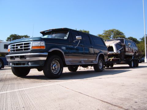 1993 Ford Bronco - A dark green Ford Bronco tows a white SUV on a trailer under a clear blue sky.