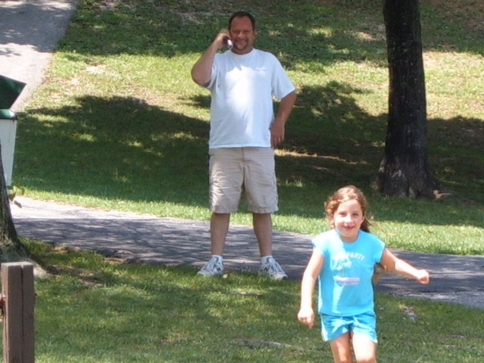 Man talking on cell phone in background, young girl running in foreground on grassy area with trees