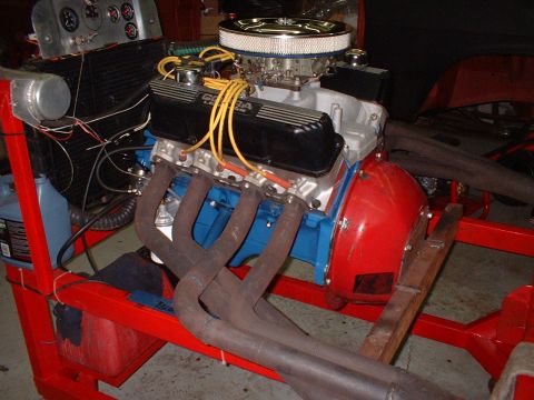 1965 Shelby Cobra - A V8 engine with black valve covers is mounted on a red stand, showing exhaust headers and a carburetor.