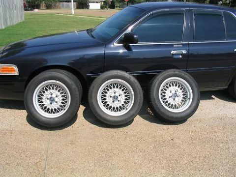 1999 Ford Crown Victoria - A dark blue sedan, possibly a 1998 Ford Crown Victoria, is parked next to three aftermarket mesh wheels.